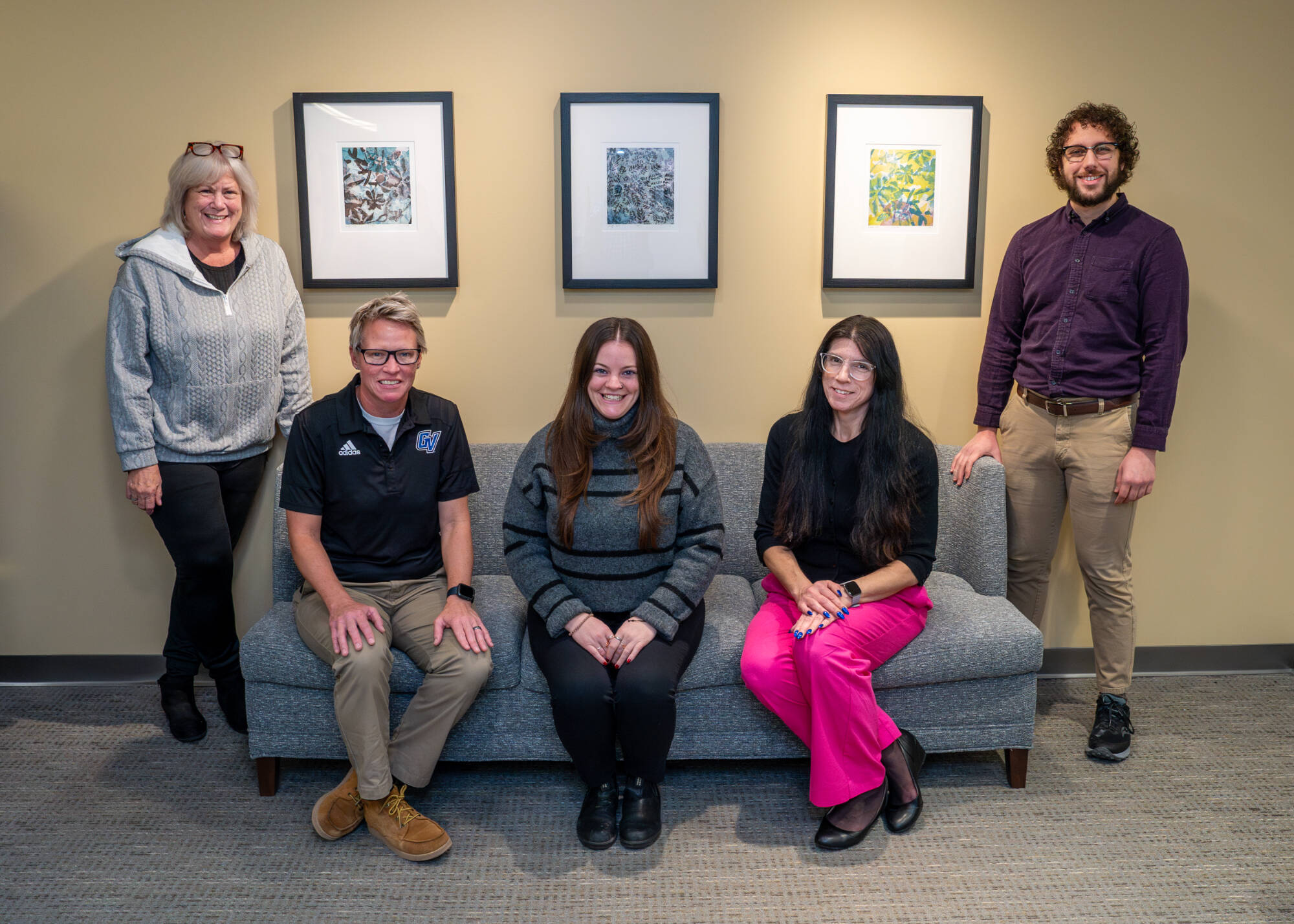 Five people in a room, three seated on a gray couch and two standing, with three framed artworks on the wall behind them.